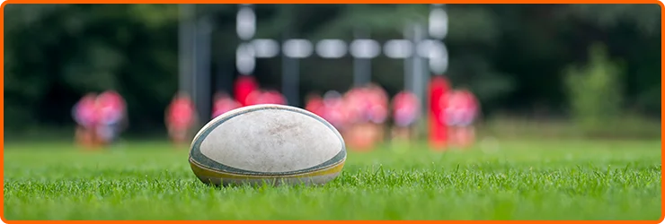 Ballon de rugby reposant sur un terrain de gazon avec des joueurs flous et des poteaux de but en arrière-plan, représentant l'entraînement professionnel, le travail d'équipe et la compétition en plein air.