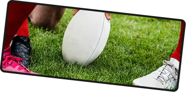 Gros plan d'un ballon de rugby posé sur de l'herbe verte entre deux joueurs se préparant au coup d'envoi, capturant l'intensité et l'anticipation avant le début du match.